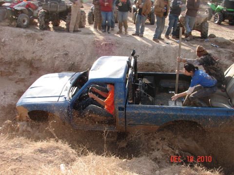 1987 Ford Bronco - A group of people attempt to free a blue pickup truck from a mud pit while ATVs and spectators observe from the embankment.