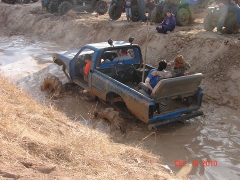 1987 Ford Bronco - Several vehicles and spectators are gathered around a blue pickup truck struggling in deep mud.