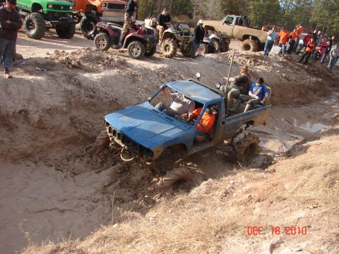 1987 Ford Bronco - A blue pickup truck navigates a muddy obstacle course, with several other vehicles and spectators visible in the background.