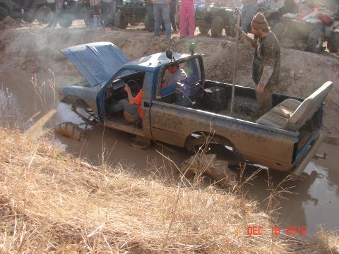 1987 Ford Bronco - A blue pickup truck is stuck in a muddy pit, with its hood open and people gathered around.