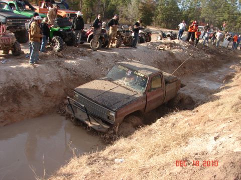 1987 Ford Bronco - A muddy pickup truck is stuck in a ditch while other vehicles and spectators watch.