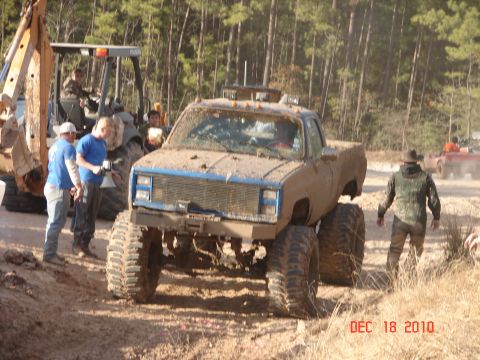 1987 Ford Bronco - Muddy pickup truck with oversized tires and a blue grille is seen in a wooded area with other vehicles.