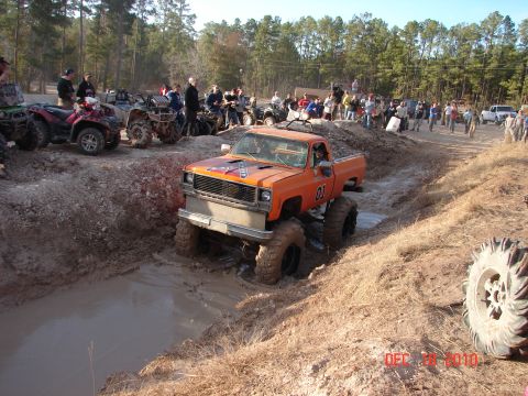 1987 Ford Bronco - A lifted orange pickup truck navigates a muddy obstacle course, while ATVs and a crowd watch from the sidelines.