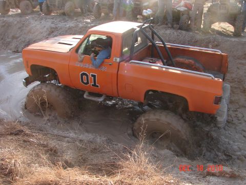 1987 Ford Bronco - An orange pickup truck navigates through a muddy terrain with large tires.