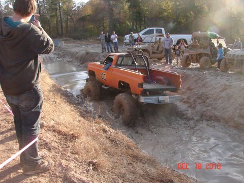 1987 Ford Bronco - A modified orange pickup truck navigates a muddy obstacle course, with spectators and other vehicles nearby.
