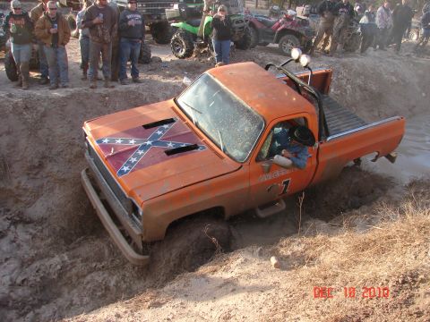 1987 Ford Bronco - A group of people watch as an orange pickup truck gets stuck in a mud pit during an off-road event.
