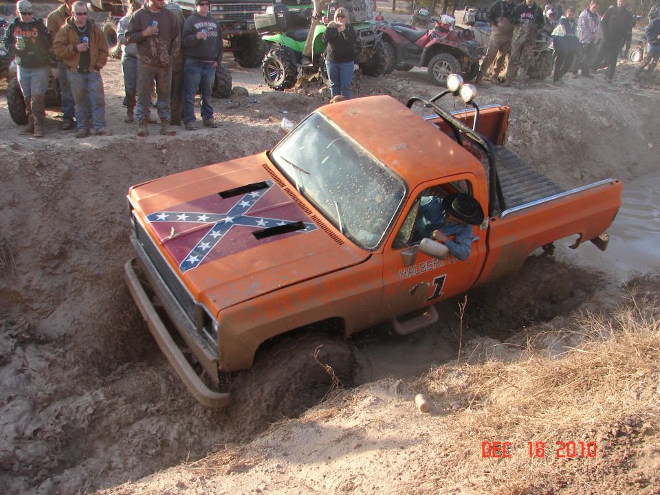 Orange pickup truck with Confederate flag on hood stuck in mud pit, people watching