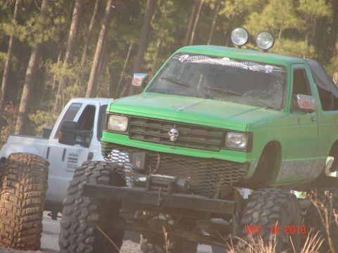1987 Ford Bronco - Two modified trucks are parked off-road in a wooded area, with the green truck in the foreground.