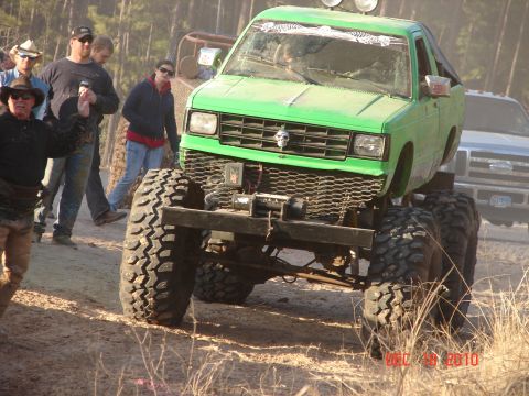 1987 Ford Bronco - A heavily modified green pickup truck with oversized tires is the focus, with other vehicles and people in the background.
