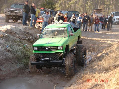 1987 Ford Bronco - A lifted green pickup truck navigates a muddy off-road course with spectators watching.