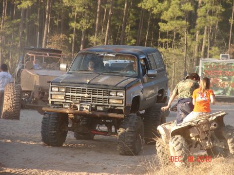 1987 Ford Bronco - A heavily modified SUV with oversized tires is seen in a muddy outdoor setting with other vehicles and people.