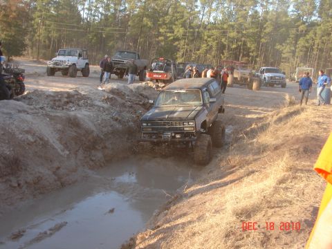 1987 Ford Bronco - Multiple off-road vehicles, including a lifted black SUV, tackle a muddy terrain during an event.