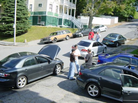 2003 Mazda 6 - Several cars are parked on a street, with people gathered around a dark sedan with its hood up.