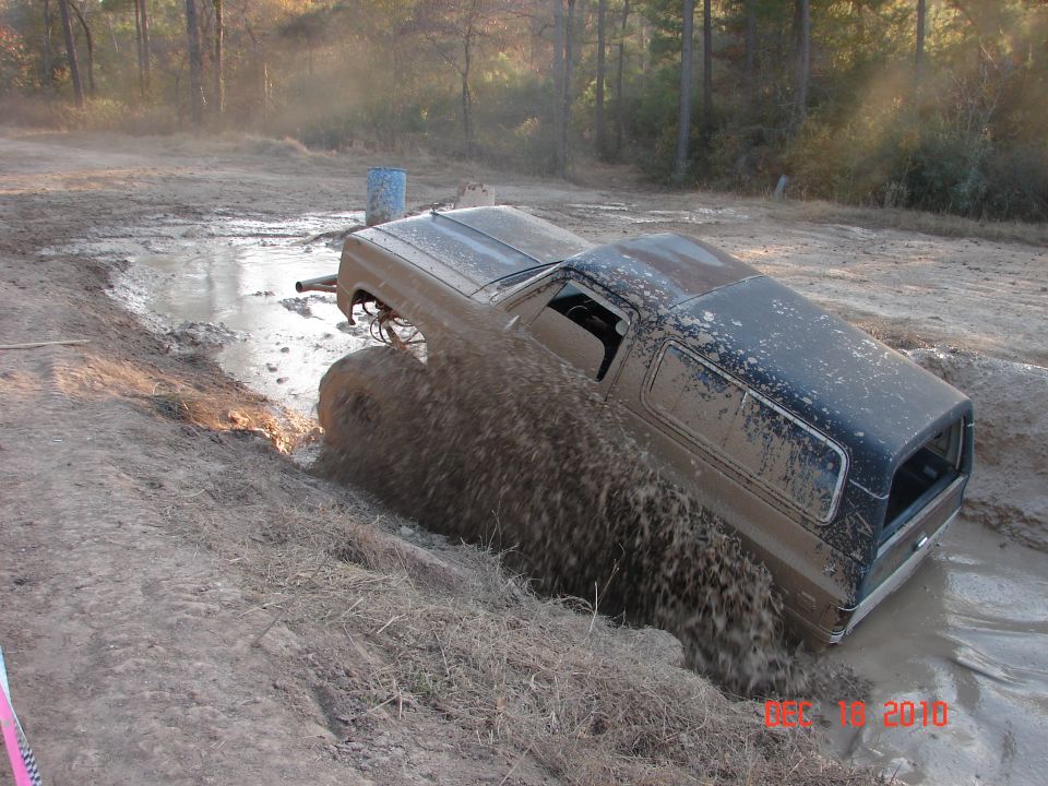 Muddy SUV stuck in a water-filled ditch, splashing water. The vehicle has a dark top and a lighter brown body