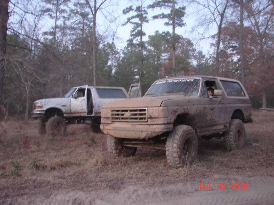 1987 Ford Bronco and another Bronco covered in mud, parked in a wooded area