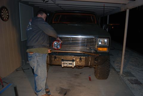 1982 Ford Bronco - A man installs a round headlight on a dark green Ford Bronco, which features an aftermarket bumper and winch.