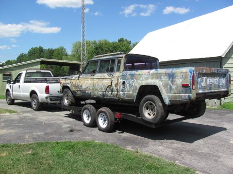 1986 Jeep Grand Wagoneer - A weathered Jeep J-10 pickup truck with camouflage paint is loaded onto a trailer behind a white Ford pickup truck.