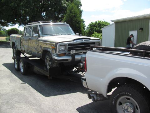 1986 Jeep Grand Wagoneer - A weathered 1986 Jeep Grand Wagoneer pickup truck is loaded onto a trailer, being towed by a white pickup truck.