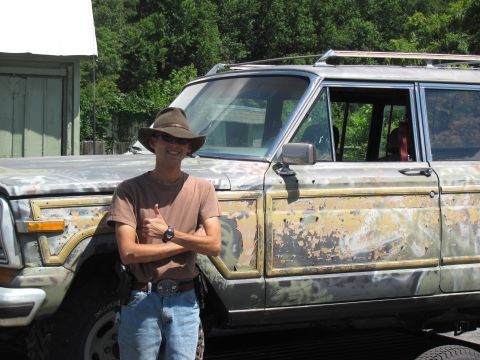 1986 Jeep Grand Wagoneer - A man poses with a heavily weathered 1986 Jeep Grand Wagoneer, showing its distinctive wood-grain paneling and roof rack.