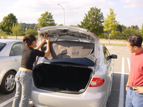 2003 Mazda 6 - A woman works on the trunk latch of a silver sedan while a man watches, with another car parked nearby.