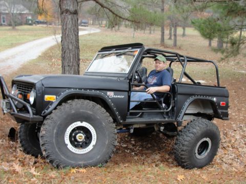 1967 Ford Mustang - A man sits in a modified black Ford Bronco, equipped with a roll cage and oversized tires, parked outdoors.