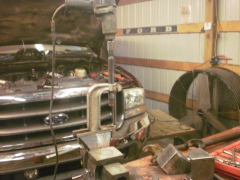 1995 Ford Bronco - A Ford truck's front end is clamped in a vise with a drill overhead, suggesting a repair or modification is underway.