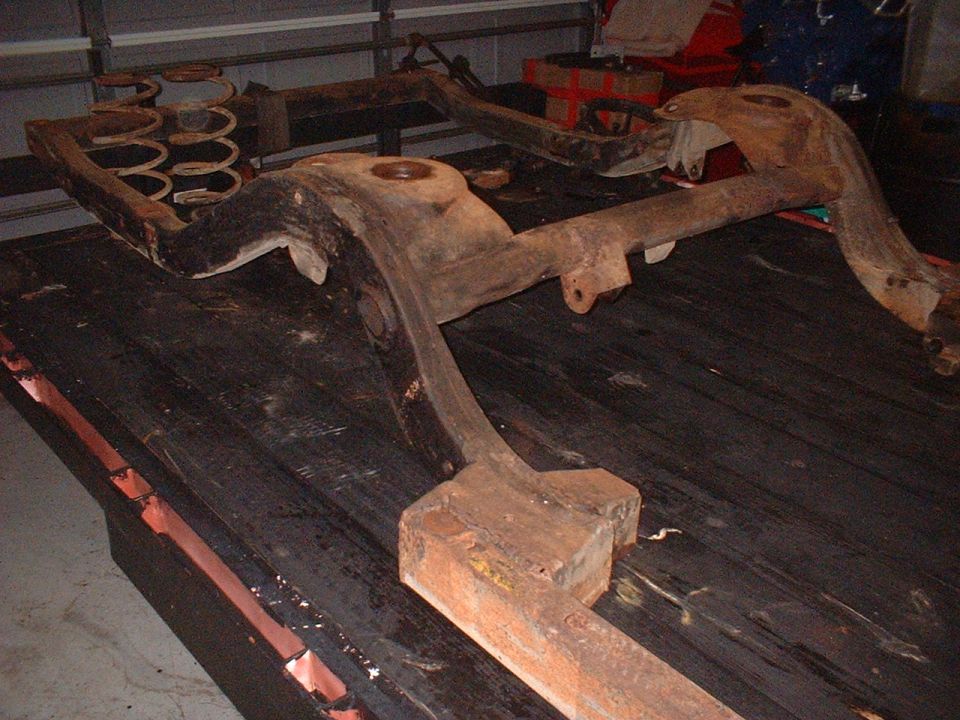Rusted vehicle subframe with coil springs, viewed from an angle, resting on a wooden surface