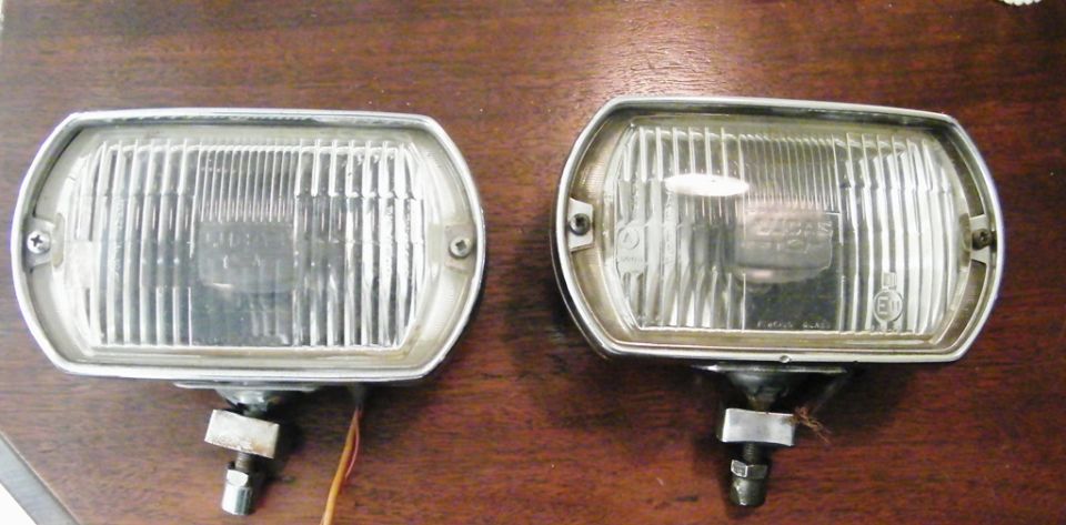 Two rectangular fog lights with chrome trim sit on a wooden surface. One has wires attached