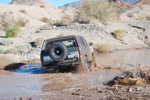 1982 Ford Bronco - A brown Ford Bronco navigates a muddy stream in a desert landscape.