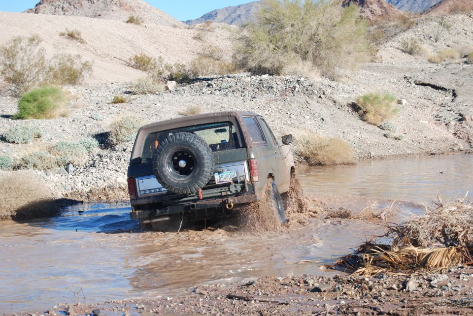 Brown Ford Bronco driving through muddy water, spare tire on rear door
