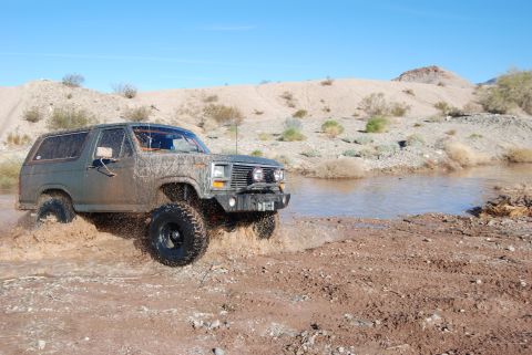 1982 Ford Bronco - A muddy green Ford Bronco navigates a water crossing in a desert landscape.