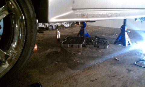 1990 Ford Mustang - The underside of a Ford Mustang GT is visible, supported by jack stands, with the oil pan removed for maintenance.