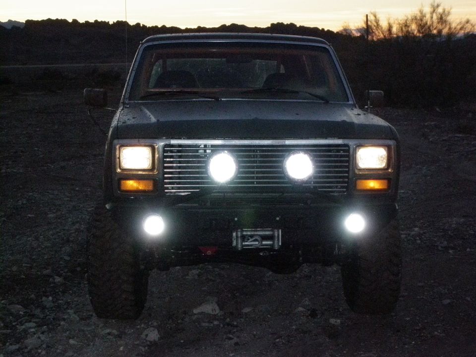 Front view of a dark-colored Ford Bronco with two large round auxiliary lights mounted on the grille and two smaller ones below the bumper. A winch is visible between the lower lights