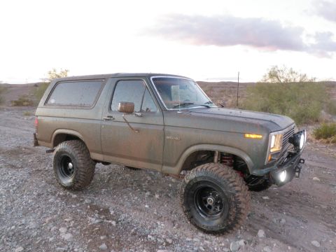 1982 Ford Bronco - This lifted Ford Bronco is shown in a three-quarter front view, parked on a rocky surface with sparse vegetation in the background.