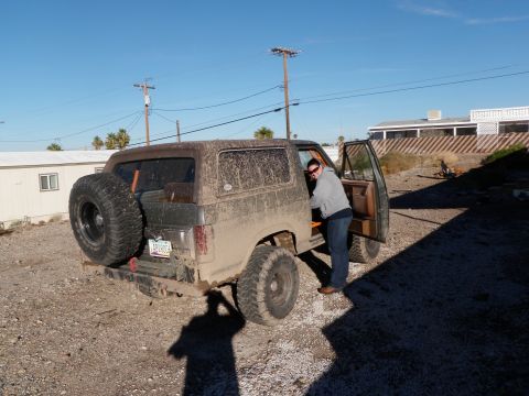 1982 Ford Bronco - A muddy Ford Bronco is parked in a lot with its driver's door open as a person gets in.