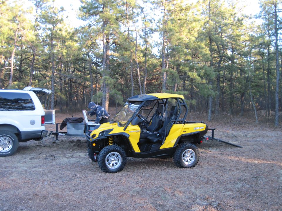 Yellow Can-Am ATV parked next to a white SUV with its trunk open. Trees in background
