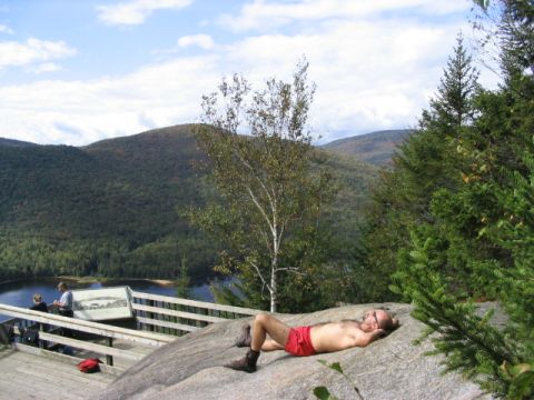 1986 Ford Bronco - A man enjoys a sunny day on a rocky outcrop with a scenic view of a lake and distant mountains.