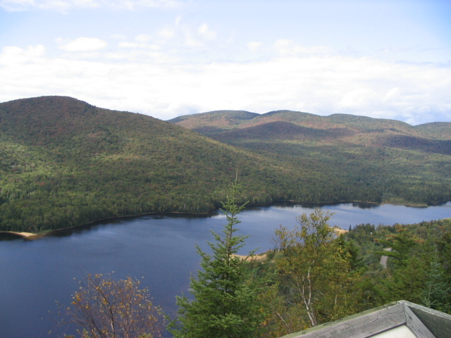 Lake surrounded by forested mountains under a cloudy sky. A road is visible in the distance
