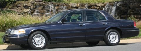 1999 Ford Crown Victoria - A dark blue 1999 Ford Crown Victoria is parked on the side of the road with a waterfall in the background.
