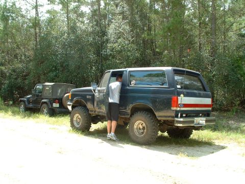 1995 Ford Bronco - Two off-road vehicles, a Ford Bronco and a Jeep Wrangler, are parked on a dirt path near trees.