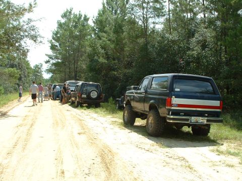 1995 Ford Bronco - Several SUVs are lined up on a dirt road next to a wooded area, with people gathered around.