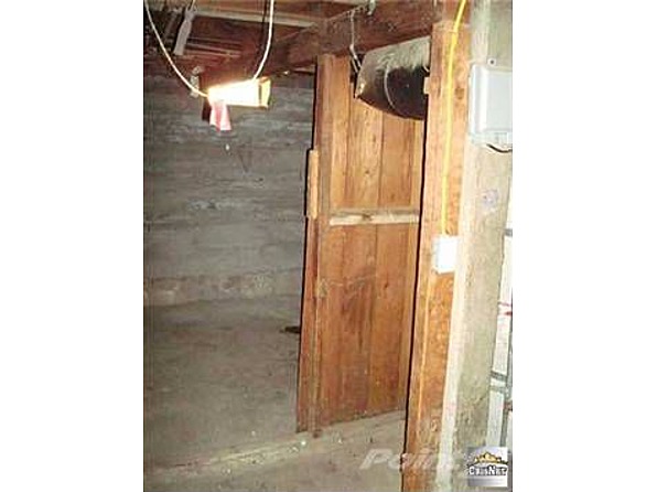 Interior of a basement with concrete floor, wooden door, and exposed ceiling beams with wiring and a light fixture