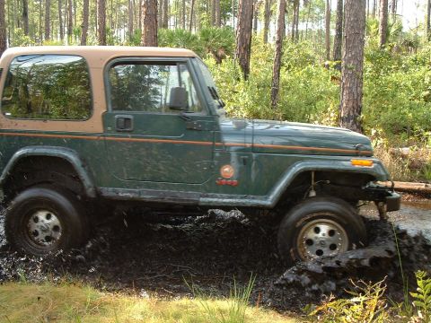 1995 Ford Bronco - A green Jeep Wrangler navigates through a muddy trail in a wooded area.