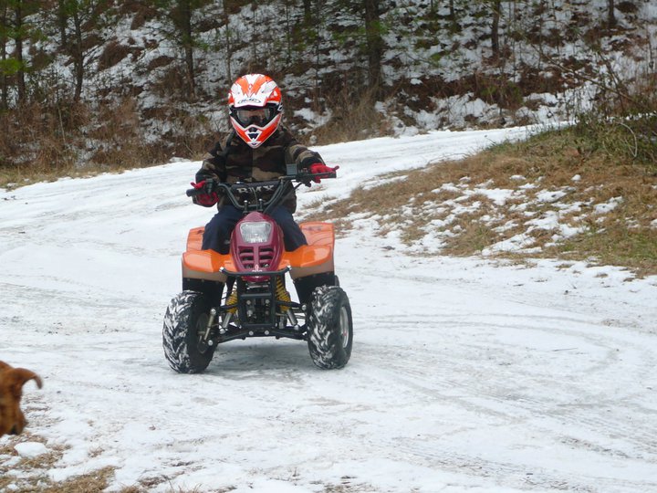 Child in motocross helmet riding an orange youth ATV on a snowy dirt road