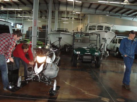 1979 Ford Bronco - Two people work on a snowmobile while a utility vehicle and boats are parked nearby in a large garage.