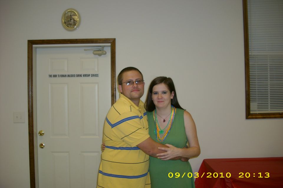 Couple posing in front of a white door with text, a clock on the wall, and a window with blinds