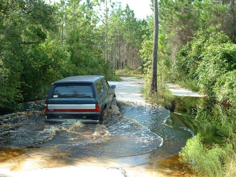1995 Ford Bronco - A blue Ford Bronco navigates a water crossing on a wooded dirt road.