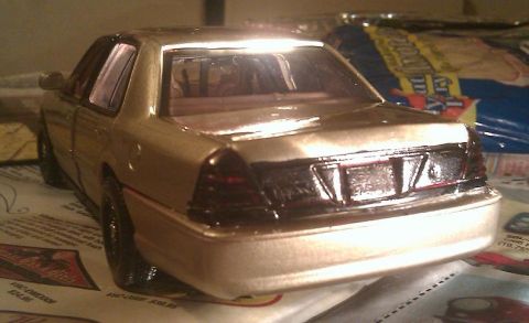 2000 Mercury Grand Marquis - A gold model car is viewed from the rear, resting on a surface with printed text and a website logo.