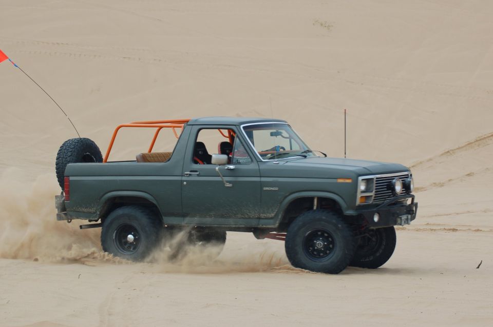 1982 Ford Bronco with orange roll cage driving through sand dunes, kicking up dust