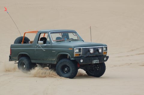1982 Ford Bronco - A green Ford Bronco with an orange roll cage navigates a sandy terrain, kicking up dust.